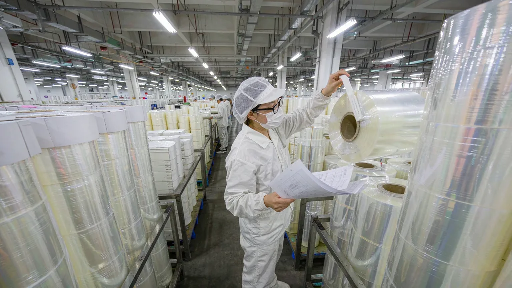 A food factory quality technician holding inspection documents next to a roll of transparent packaging film in a warehouse aisle